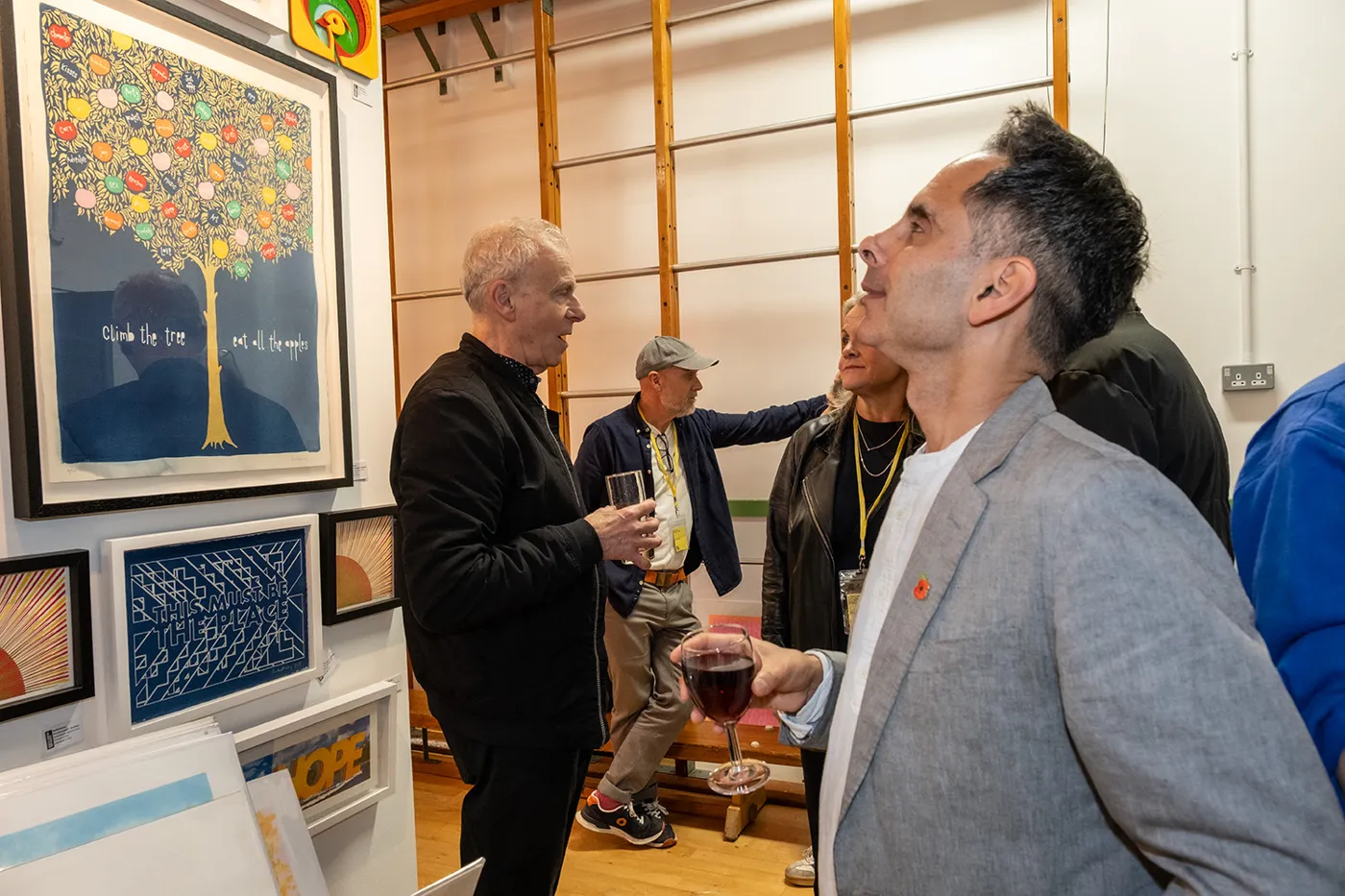 Several men talking, some holding glasses of wine, beside walls of framed prints