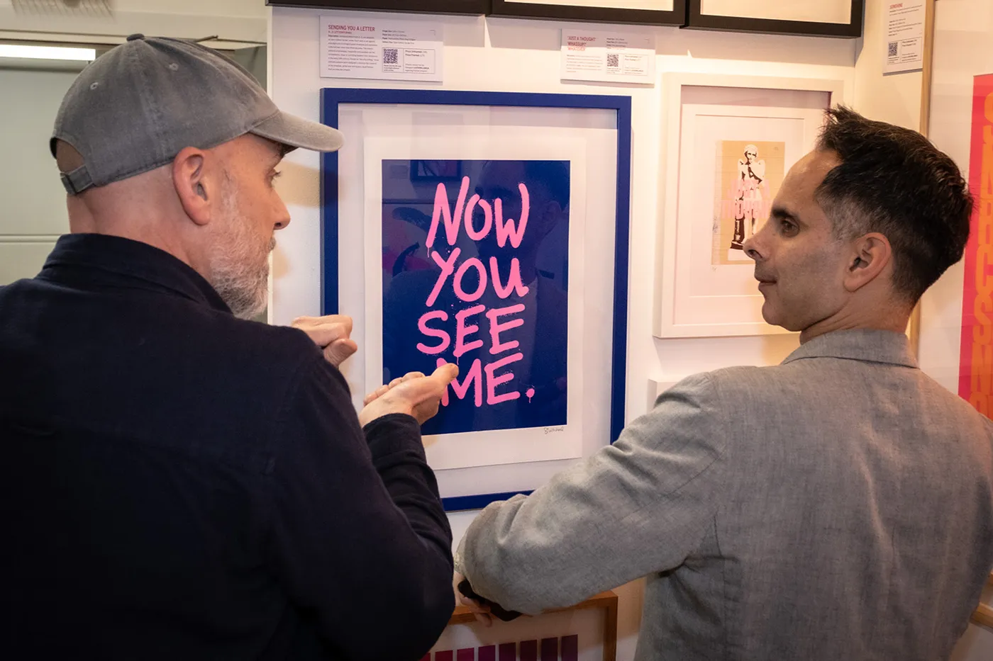 Two men in front of a framed pink and blue print reading Now You See Me