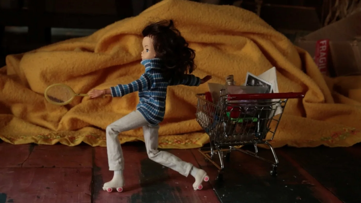 Stop-motion still of a doll in a striped jumper running past a toy shopping trolley in front of a yellow blanket