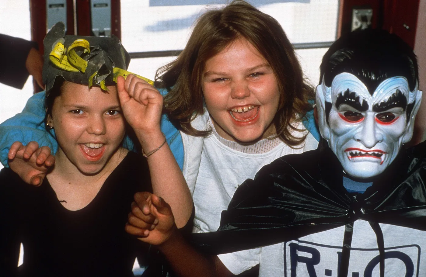 Three children laughing at the camera, one in a cardboard hat and one in a vampire mask and cape over a Rio T-shirt