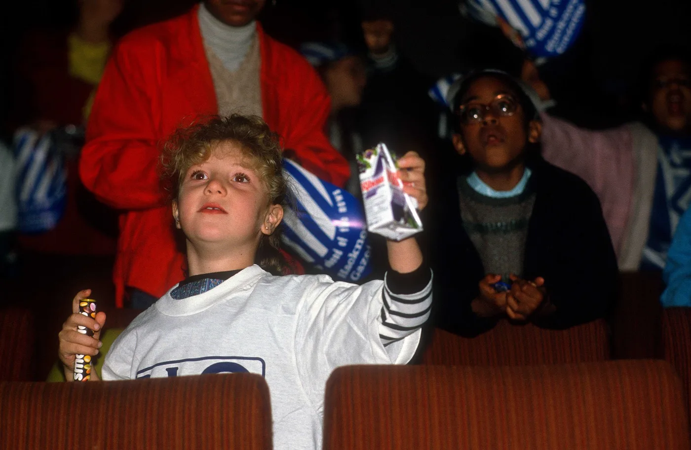 A boy in a Rio T-shirt holding up a drinks carton in a row of red cinema seats, with other children behind