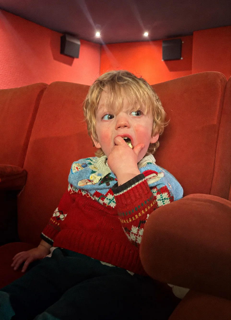 A small child in a red jumper eating popcorn in a red cinema seat
